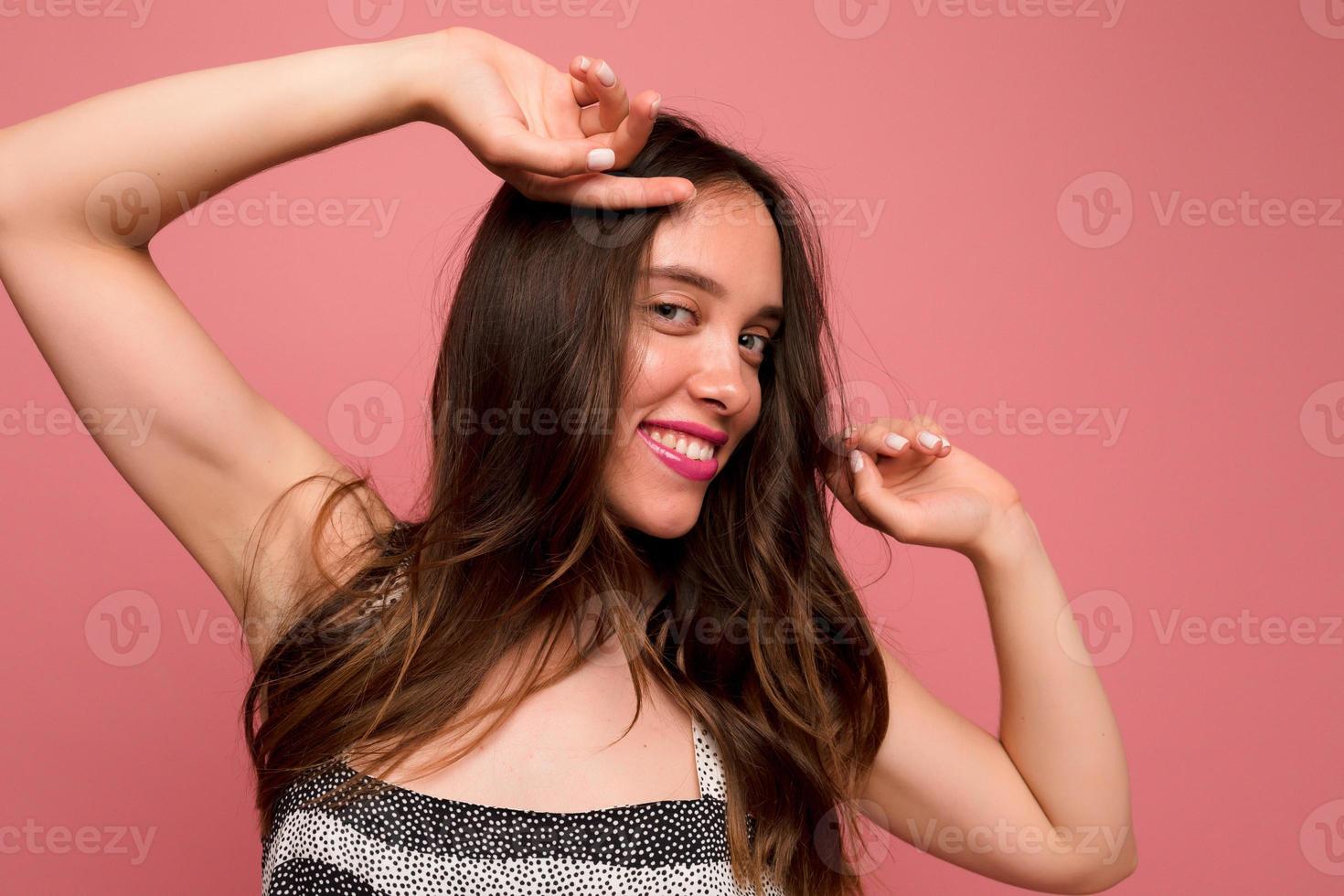 portrait en gros plan d'une jolie fille mignonne avec des cheveux bruns et un sourire merveilleux avec des lèvres roses touche les cheveux et regarde la caméra sur fond rose photo