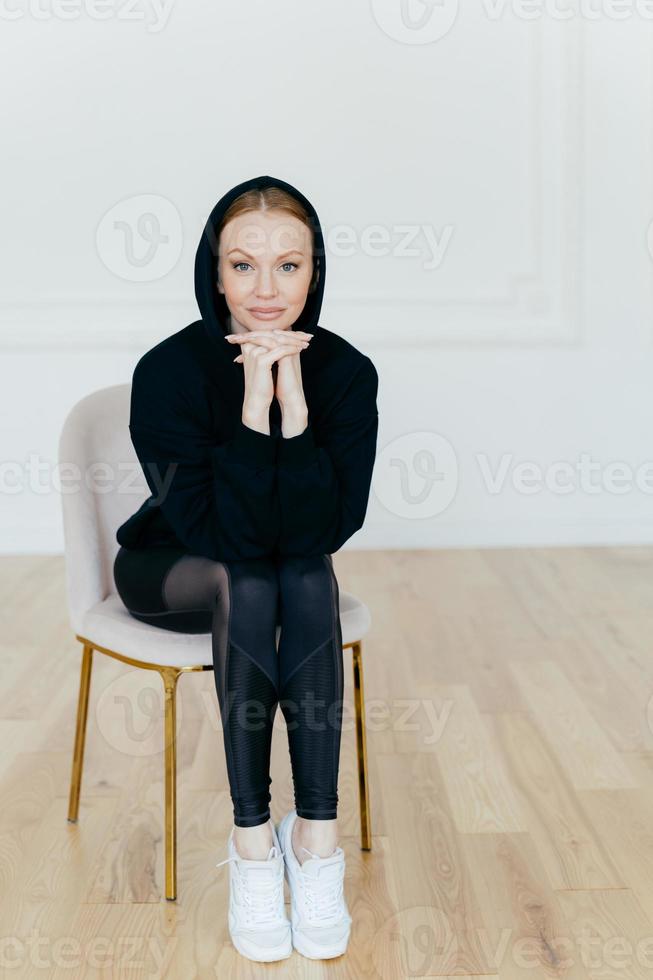 photo verticale d'une belle femme maquillée, garde les mains sous le menton, porte un sweat à capuche, des leggings et des chaussures de sport blanches, a des jambes fines, pose sur une chaise dans une pièce vide, prête pour l'entraînement sportif