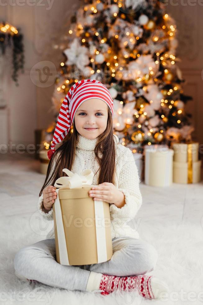 portrait vertical d'un beau petit enfant à l'air agréable porte un pull et des chaussettes tricotés, est assis les jambes croisées avec un cadeau, a le désir de l'envelopper, étant dans le salon près de l'arbre du nouvel an décoré photo