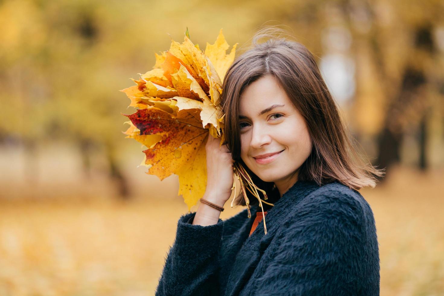 jolie jeune femme européenne souriante se promène dans le parc, profite d'une journée ensoleillée en automne, porte des feuilles jaunes, porte un manteau, pose sur un arrière-plan flou. concept de personnes, de loisirs et de saison. photo