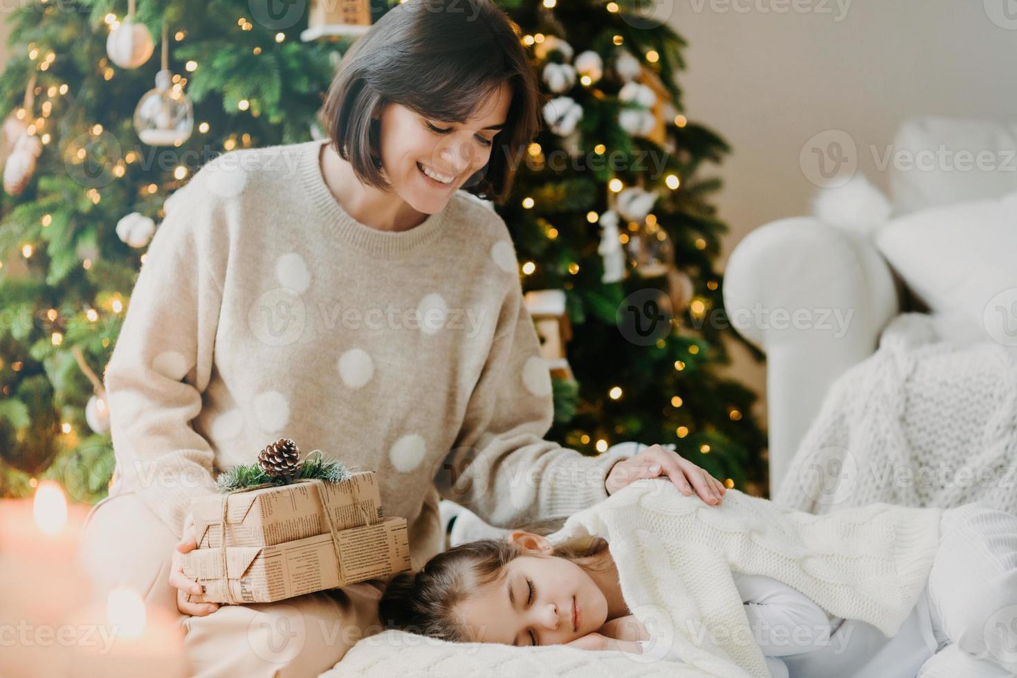 j'ai un petit secret pour toi. belle mère et fille célèbrent le nouvel an à la maison. maman pose près d'une fille endormie avec des cadeaux contre l'arbre du nouvel an décoré. concept de joyeuses vacances d'hiver photo