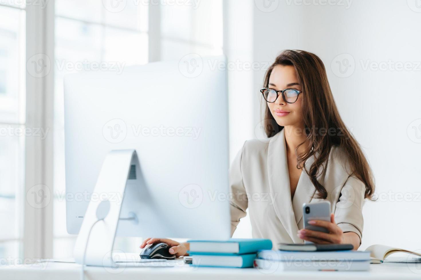 jolie femme entrepreneur aux longs cheveux noirs, travaille sur ordinateur, développe une stratégie de campagne publicitaire, pose dans un bureau de coworking moderne, utilise un téléphone portable pour discuter en ligne, porte des lunettes photo