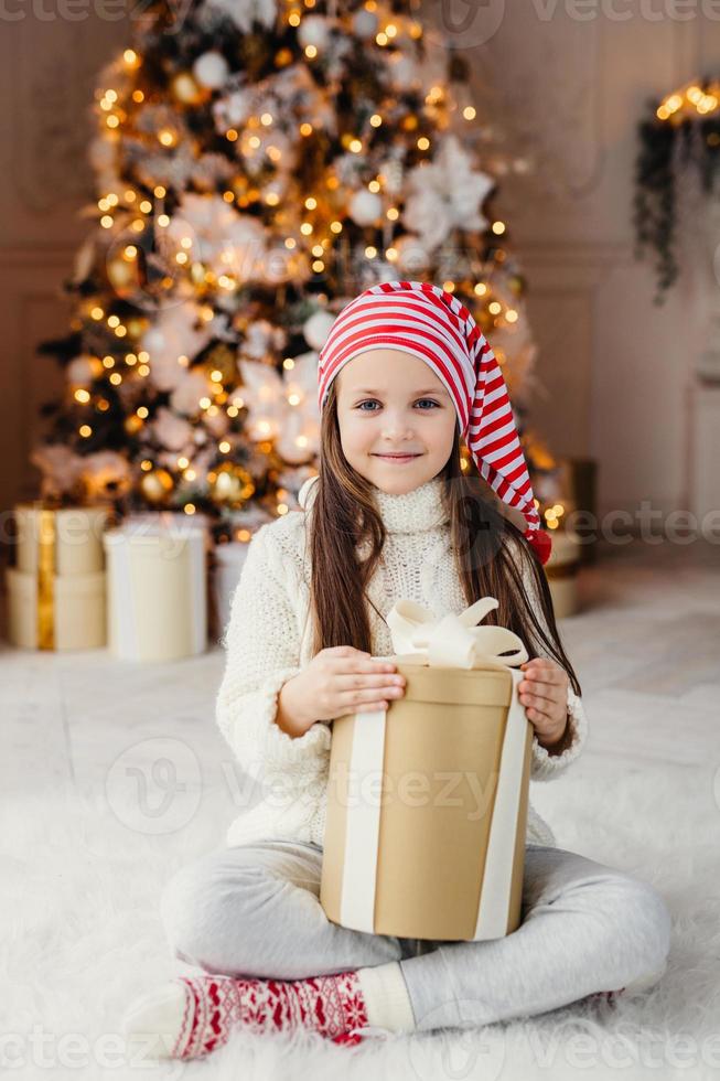 portrait vertical d'un beau petit enfant à l'air agréable porte un pull et des chaussettes tricotés, est assis les jambes croisées avec un cadeau, a le désir de l'envelopper, étant dans le salon près de l'arbre du nouvel an décoré photo