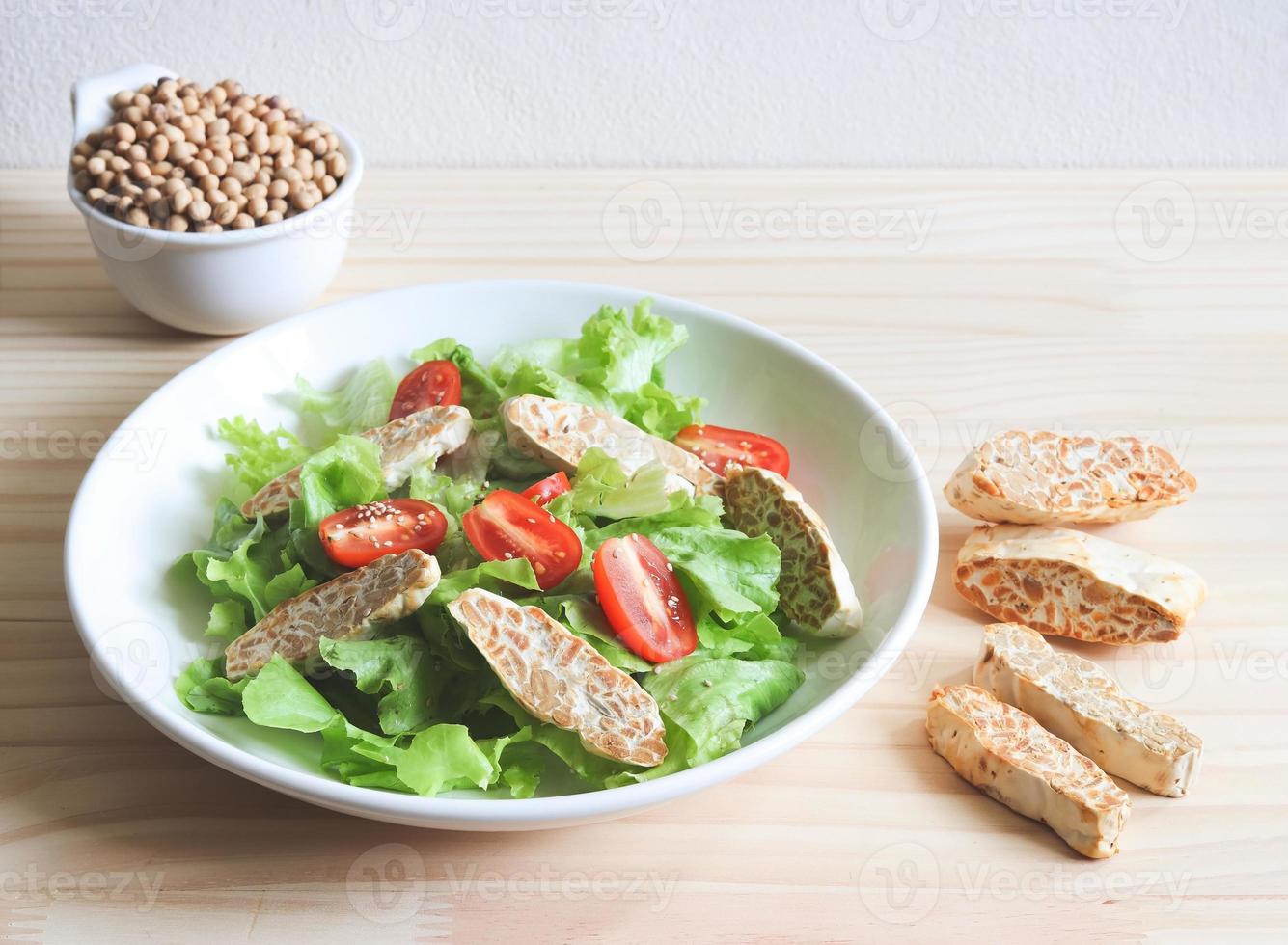 salade de tempeh ou de tempeh avec tomate et légume vert dans une assiette blanche et un bol de fèves de soja sur une table en bois. photo