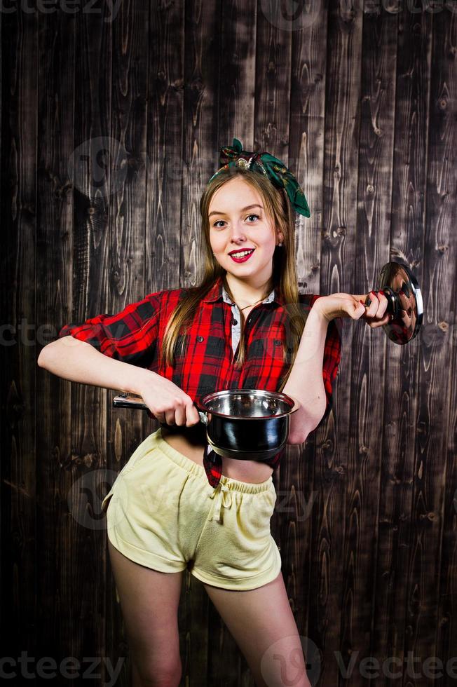 jeune femme au foyer drôle en chemise à carreaux et short jaune pin up style avec casserole sur fond en bois. photo