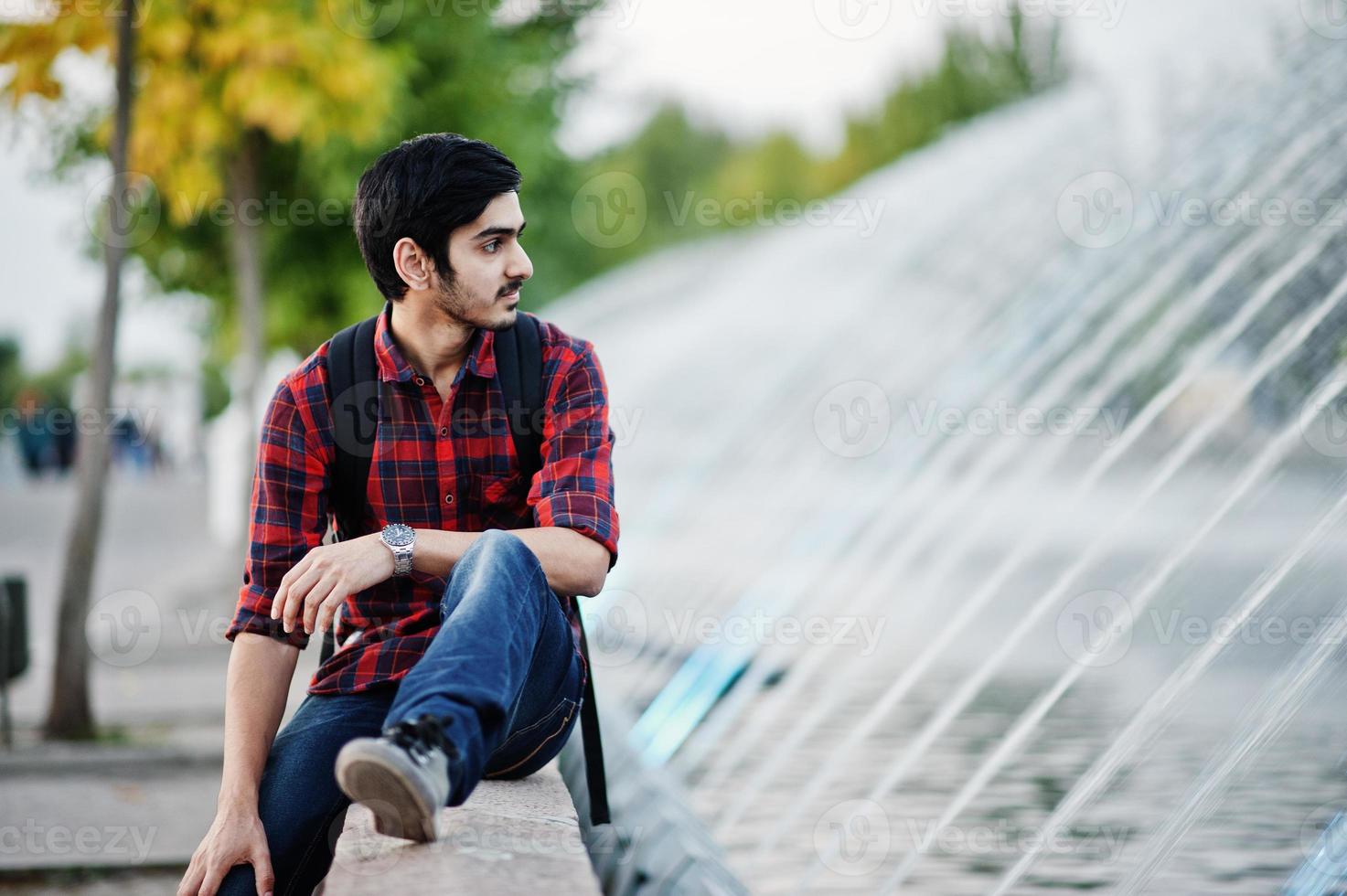 jeune étudiant indien en chemise à carreaux et jeans avec sac à dos posé sur la ville du soir contre les fontaines. photo