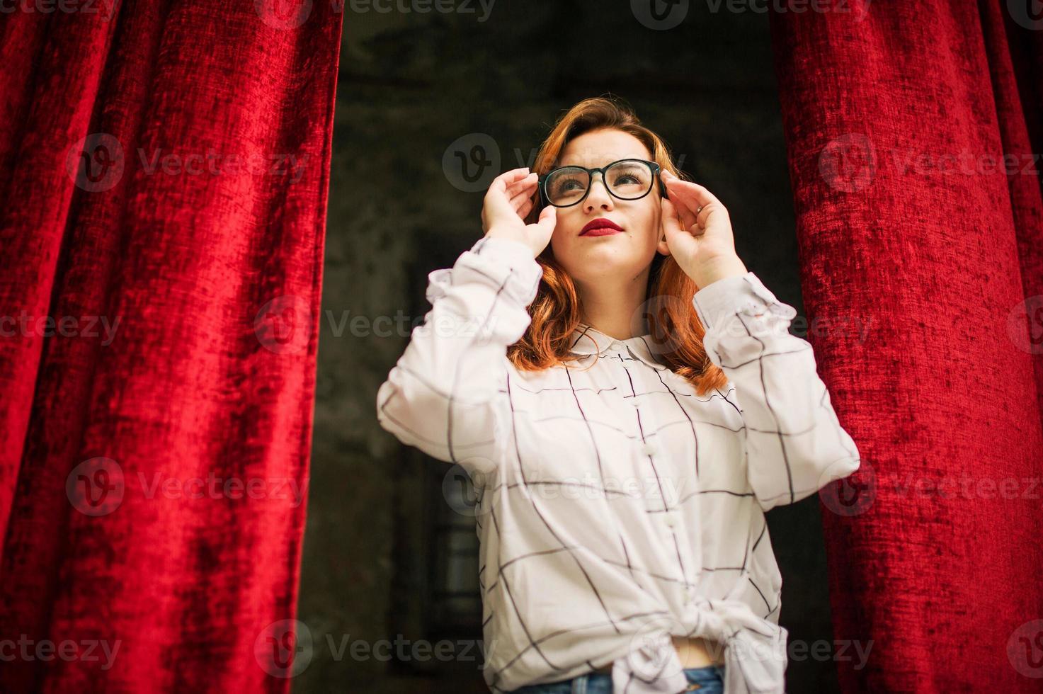 jolie femme rousse à lunettes, porter sur un chemisier blanc posant à l'arche de rideaux rouges ouverts. photo