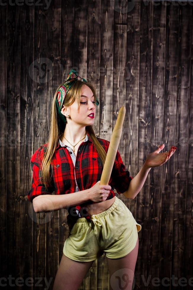 jeune femme au foyer drôle en chemise à carreaux et short jaune style pin up avec rouleau à pâtisserie de cuisine sur fond en bois. photo