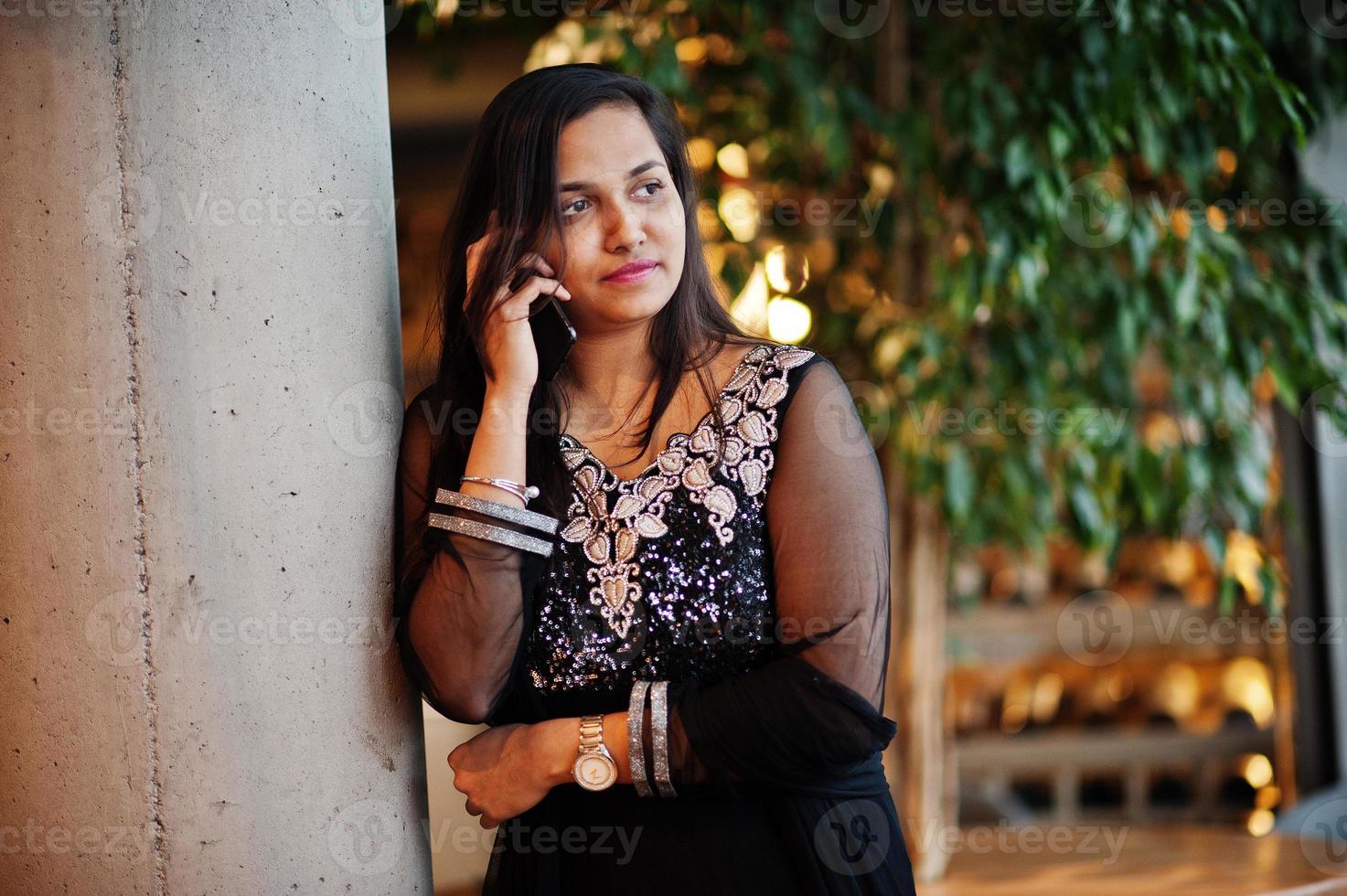 jolie fille indienne en robe saree noire posée au restaurant avec un téléphone portable à portée de main. photo