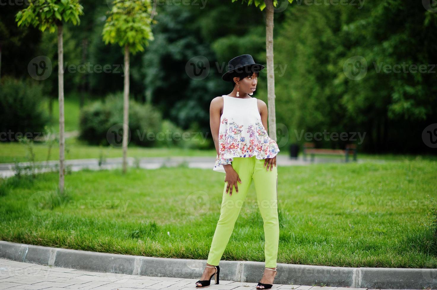 incroyable femme modèle afro-américaine en pantalon vert et chapeau noir posé au parc. photo