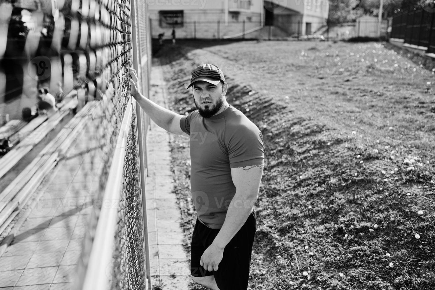 jeune homme musclé barbu brutal portant une chemise rouge, un short et une casquette au stade. photo