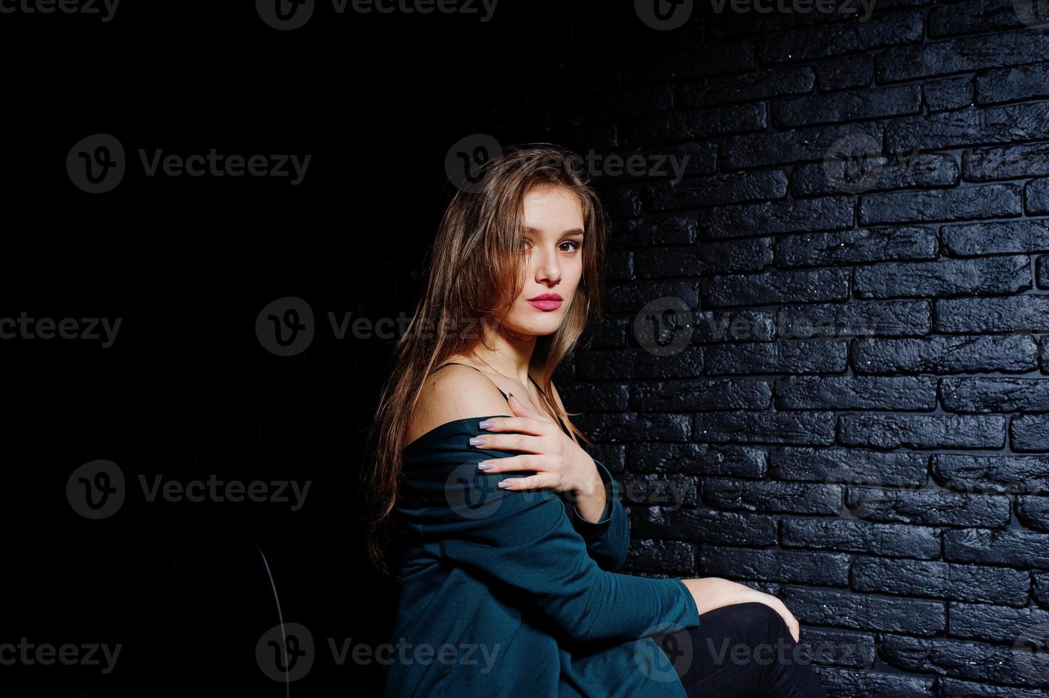 belle fille brune portant une veste noire et verte, assise et posant sur une chaise au studio contre un mur de briques sombres. portrait de modèle de studio. photo