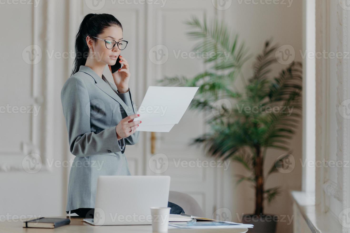 une employée brune sérieuse se tient près du bureau, tient des documents papier, parle via un smartphone, porte des vêtements et des lunettes formels, discute de la stratégie commerciale avec son partenaire, organise une réunion photo