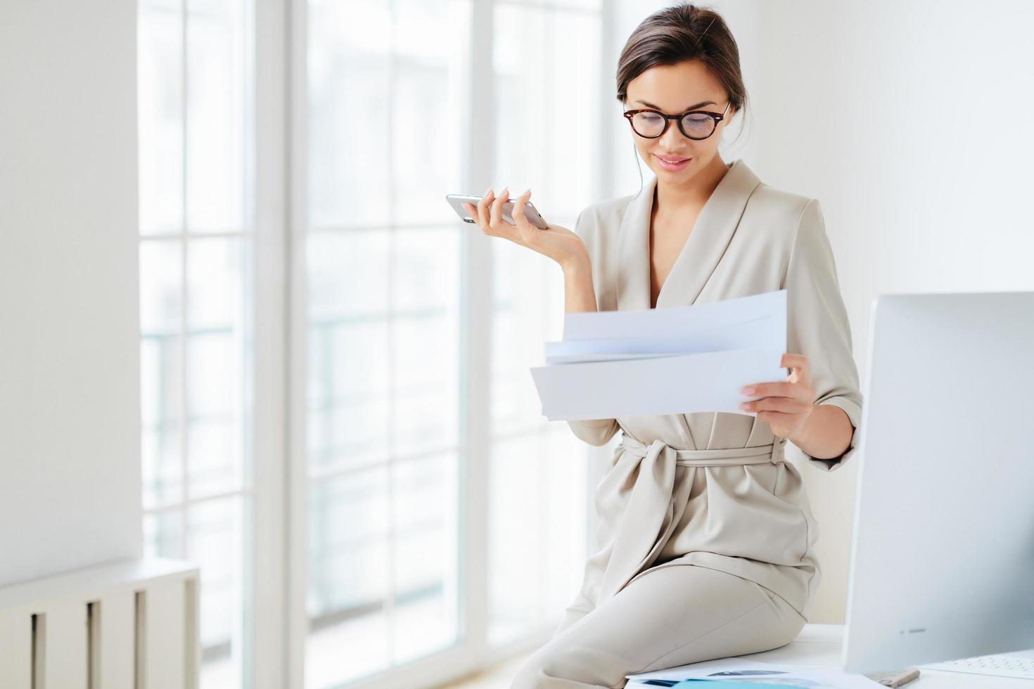 photo d'une belle femme entrepreneur en costume formel, concentrée sur un smartphone, vérifie les informations des documents papier, tient le téléphone, va chercher des nouvelles financières en ligne. l'avocat se prépare pour le briefing