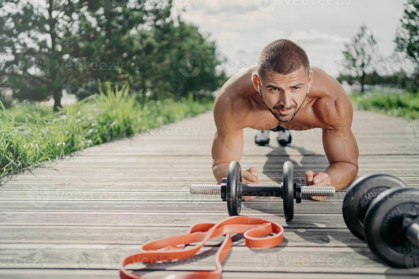 un bel homme fort et motivé se tient dans une pose de planche, fait des exercices abdominaux, pose près d'un équipement de sport, écoute de la musique dans des écouteurs sans fil, s'entraîne en plein air, regarde avec une expression sérieuse photo