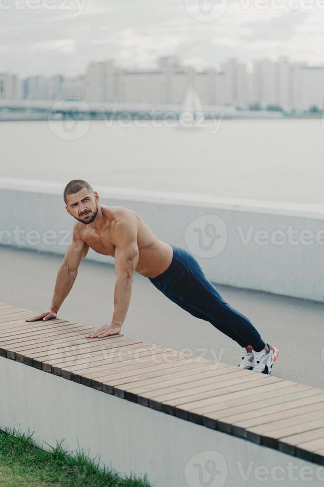 photo en extérieur d'un homme musclé déterminé s'échauffe avant l'entraînement se tient en pose de planche a le torse nu le corps musclé aime faire de l'exercice en plein air porte des vêtements de sport s'entraîne pour maintenir sa vitalité