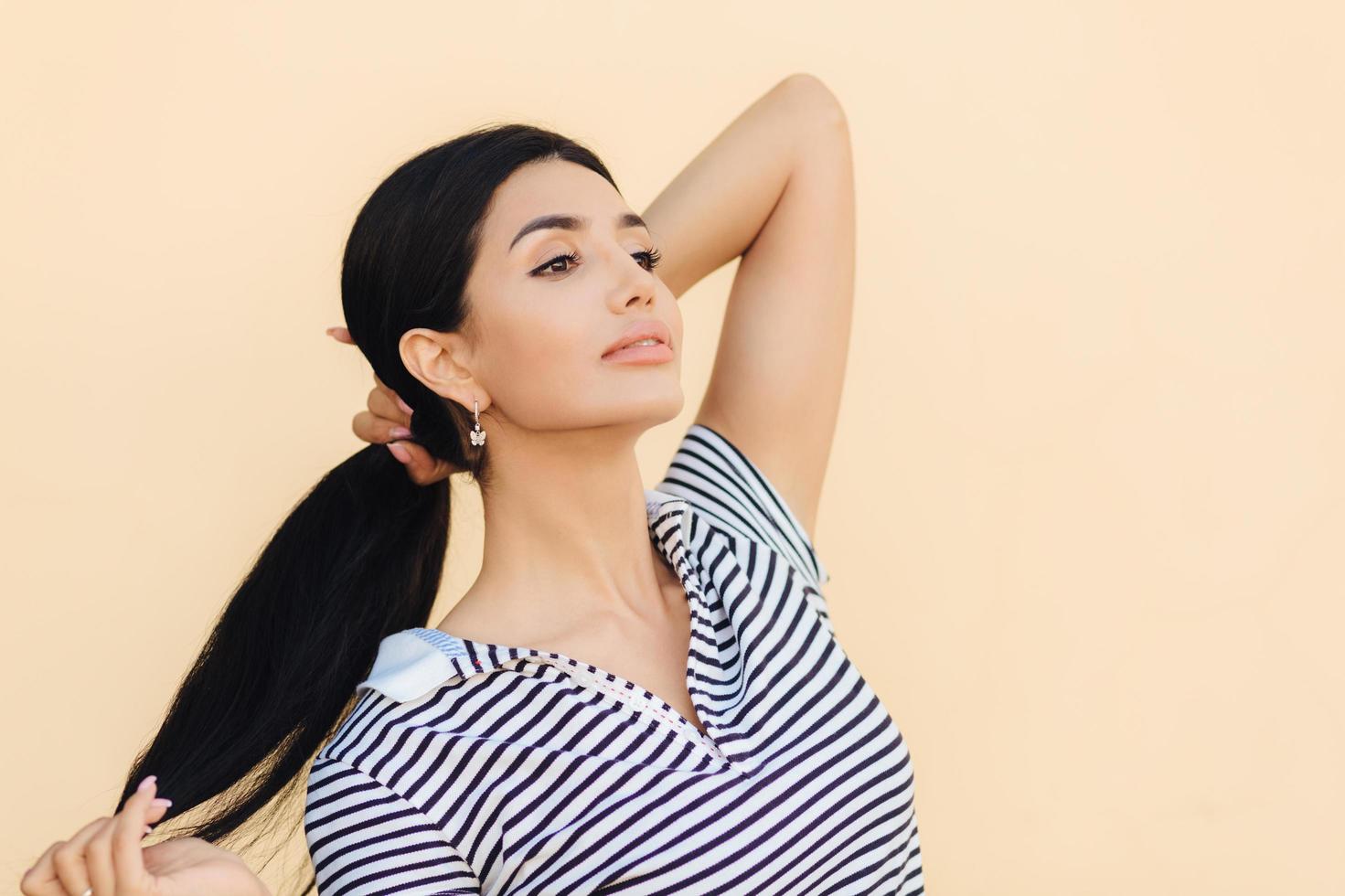 photo de côté d'une belle femme brune qui garde les cheveux en queue de cheval, regarde avec confiance au loin, pense à quelque chose, montre ses cheveux luxueux, se tient à l'intérieur contre un mur de studio lumineux