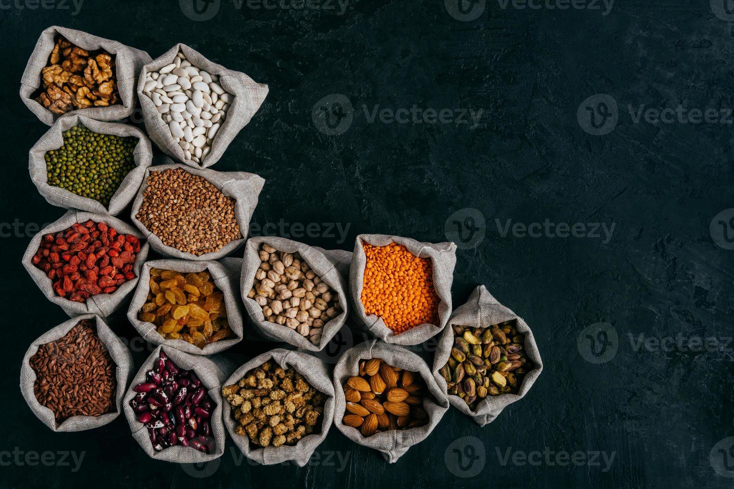 photo horizontale de petits sacs remplis d'amandes, de noix, de raisins secs, de pois chiches, de haricots rouges, de goji rouge, isolés sur fond sombre, espace vide pour votre publicité