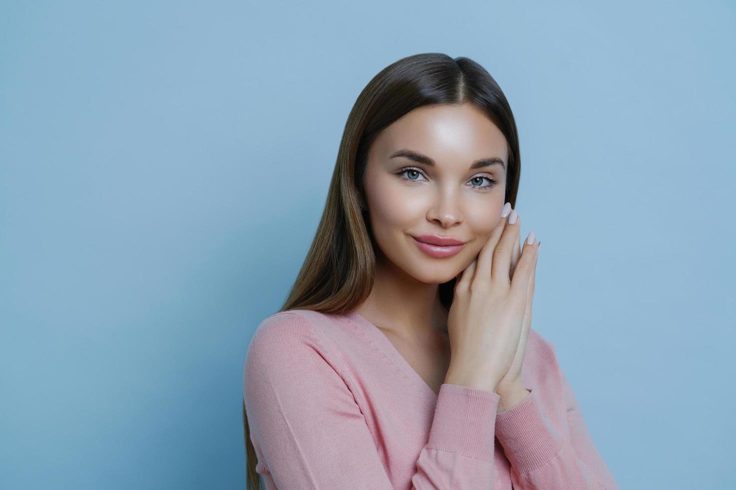 portrait en gros plan d'une jolie femme brune ravie regarde directement la caméra, garde les mains près du visage, écoute les conseils du cosmétologue sur la façon de prendre soin de la peau, les modèles à l'intérieur contre le mur bleu photo