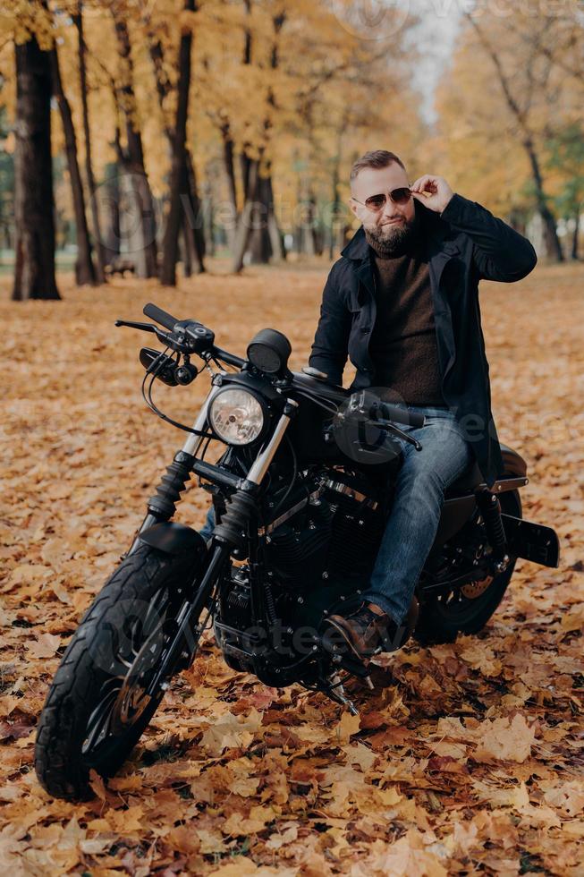 image verticale d'un motard réfléchi porte des lunettes de soleil, fait de la moto dans le parc, pose dans la rue, porte des vêtements élégants, aime le temps libre et les loisirs, admire la nature, profite du temps automnal ensoleillé photo