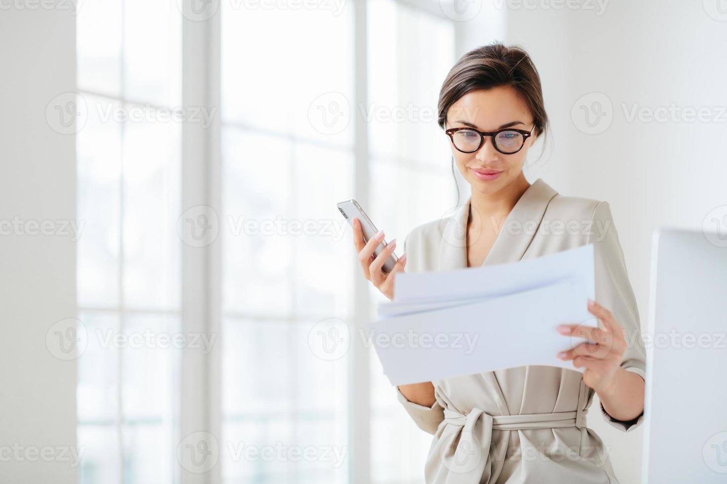 une employée satisfaite concentrée sur les documents, lit attentivement le rapport préparé, tient un smartphone moderne, porte des lunettes transparentes et des poses de costume d'affaires sur l'intérieur du bureau vérifie le revenu mensuel photo