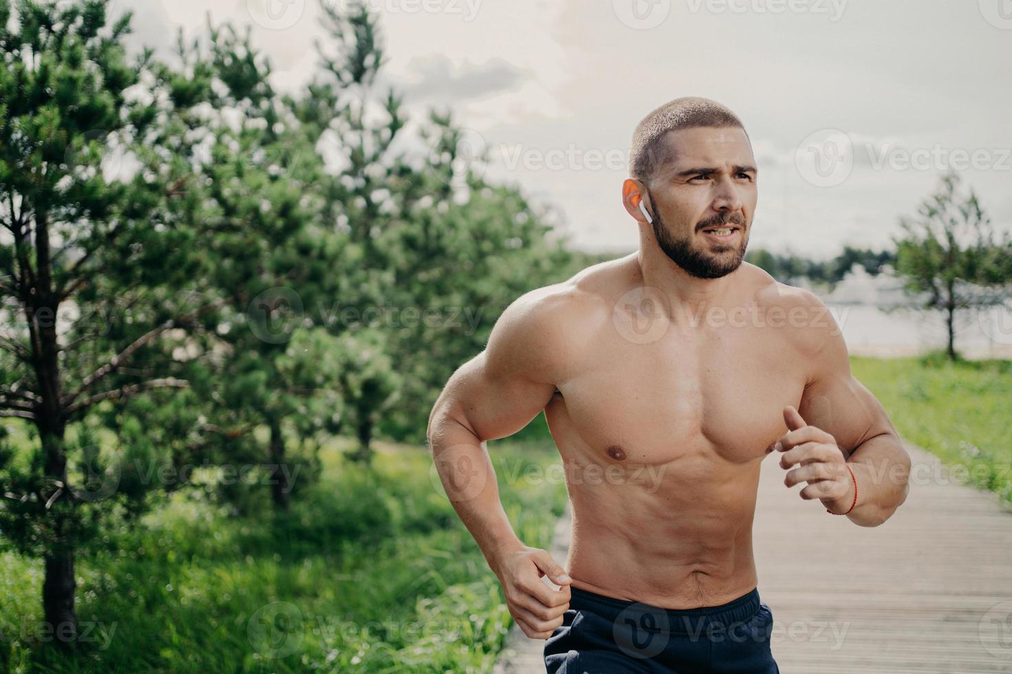 un homme athlétique sportif court à l'extérieur avec un torse nu, mène une vie saine, s'entraîne le matin, écoute de la musique dans des écouteurs sans fil, a un corps musclé, respire de l'air frais. activités de santé photo