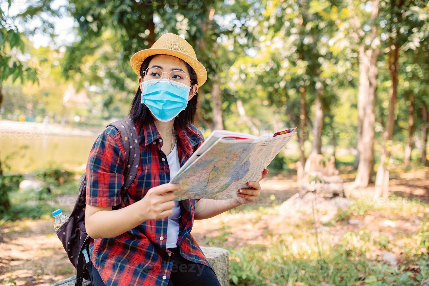 femme asiatique tourisme voyageant dans un magnifique parc verdoyant portant une protection avec un masque avec appareil photo et sac à dos et tenant sa carte, femmes asiatiques et regardant dehors pendant s'asseoir sur une chaise en pierre