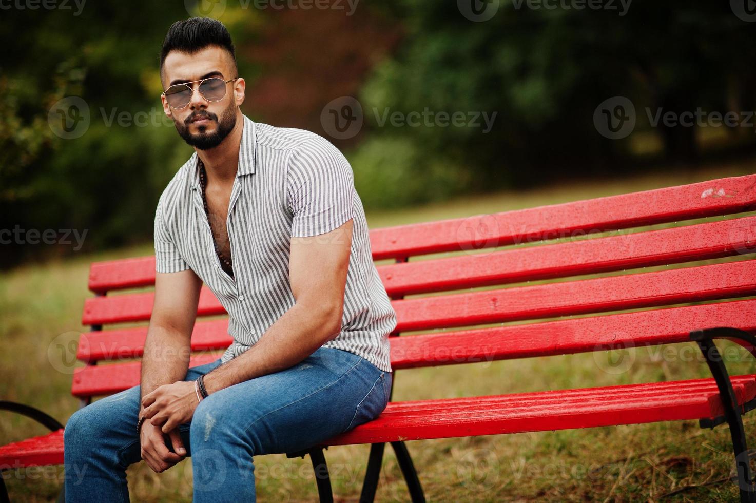 grand homme à la barbe arabe à la mode portant une chemise, un jean et des lunettes de soleil assis sur un banc rouge au parc. photo