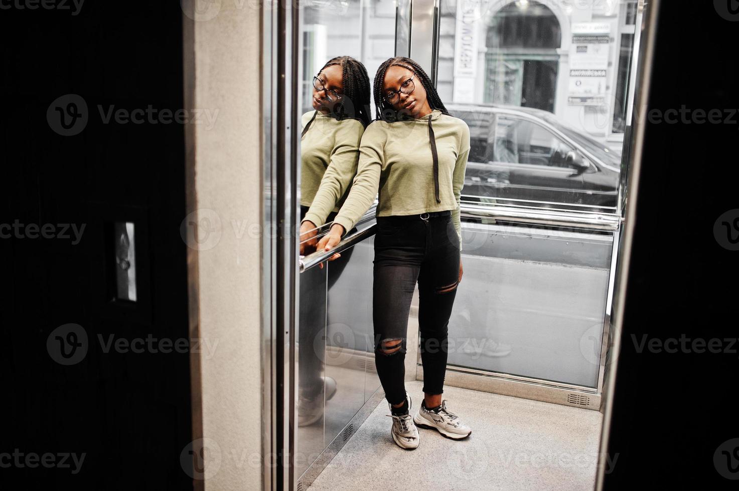 portrait d'une jeune femme positive à la peau foncée portant un sweat à capuche vert et des lunettes debout à l'ascenseur. photo