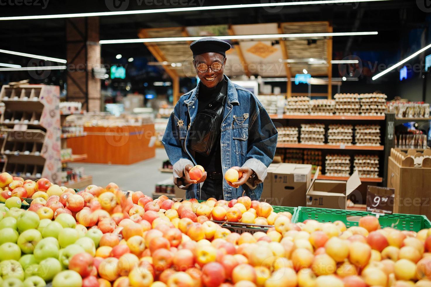 élégant homme afro-américain décontracté à la veste en jean et au béret noir vérifiant les fruits de pomme dans la section bio du supermarché. photo