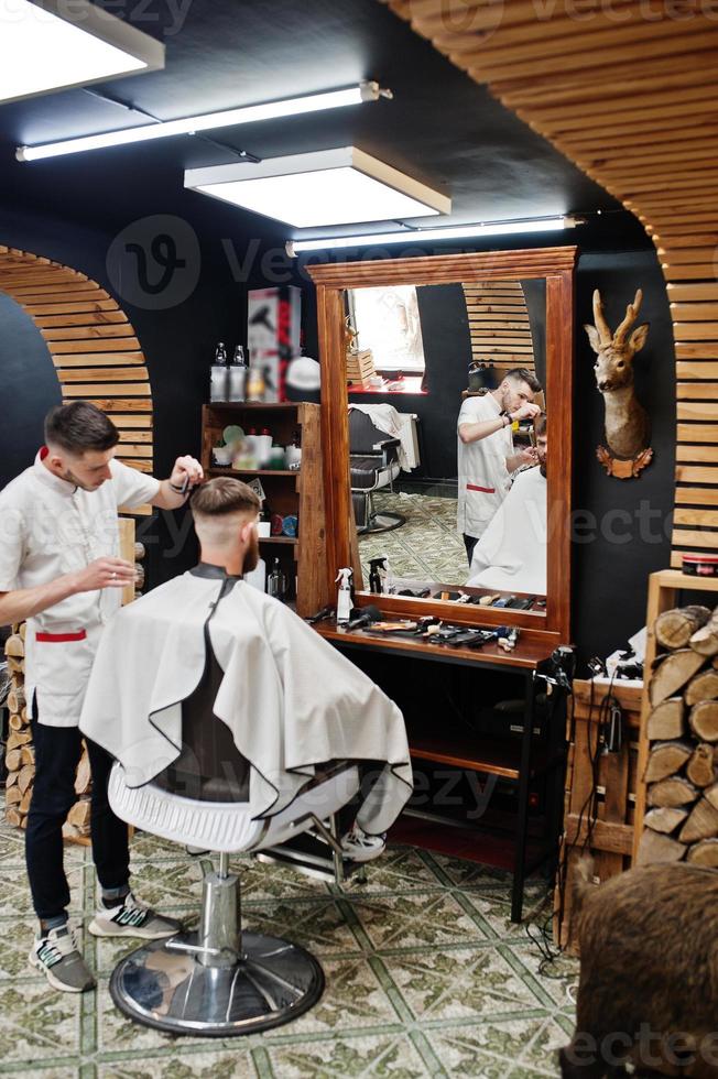 jeune homme barbu se coupe les cheveux par un coiffeur assis sur une chaise au salon de coiffure. âme de barbier. photo