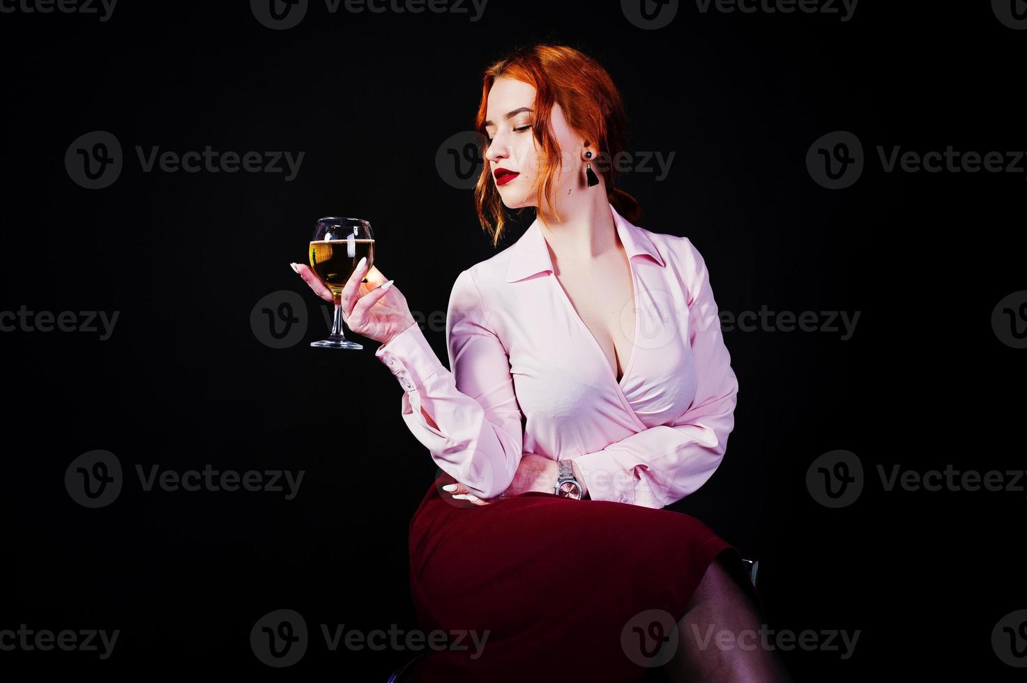 magnifique fille aux cheveux rouges en chemisier rose et jupe rouge avec un verre de vin à portée de main isolé sur noir. photo