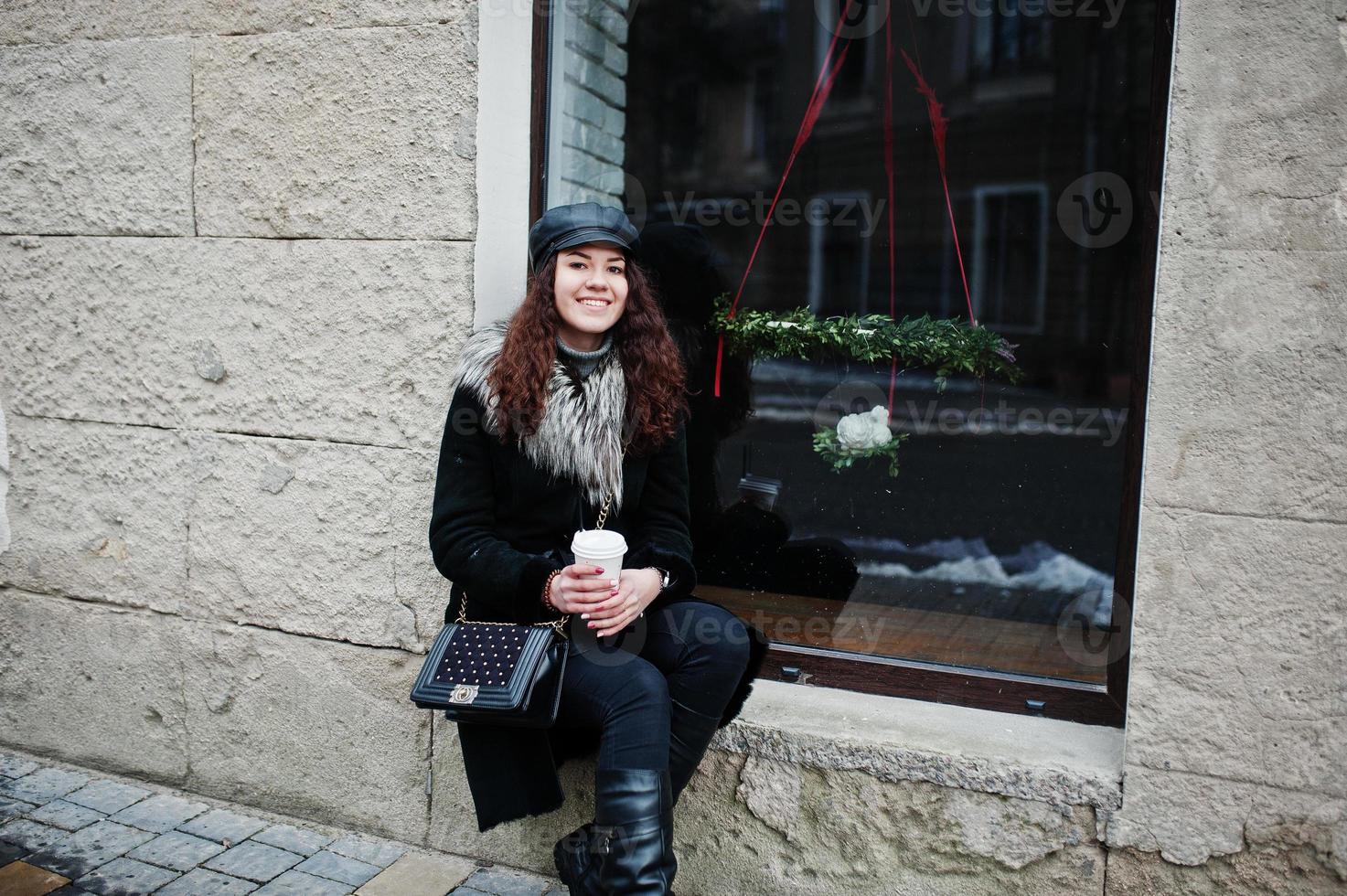 fille mexicaine bouclée en casquette en cuir et tasse de café en plastique à portée de main marchant dans les rues de la ville. photo