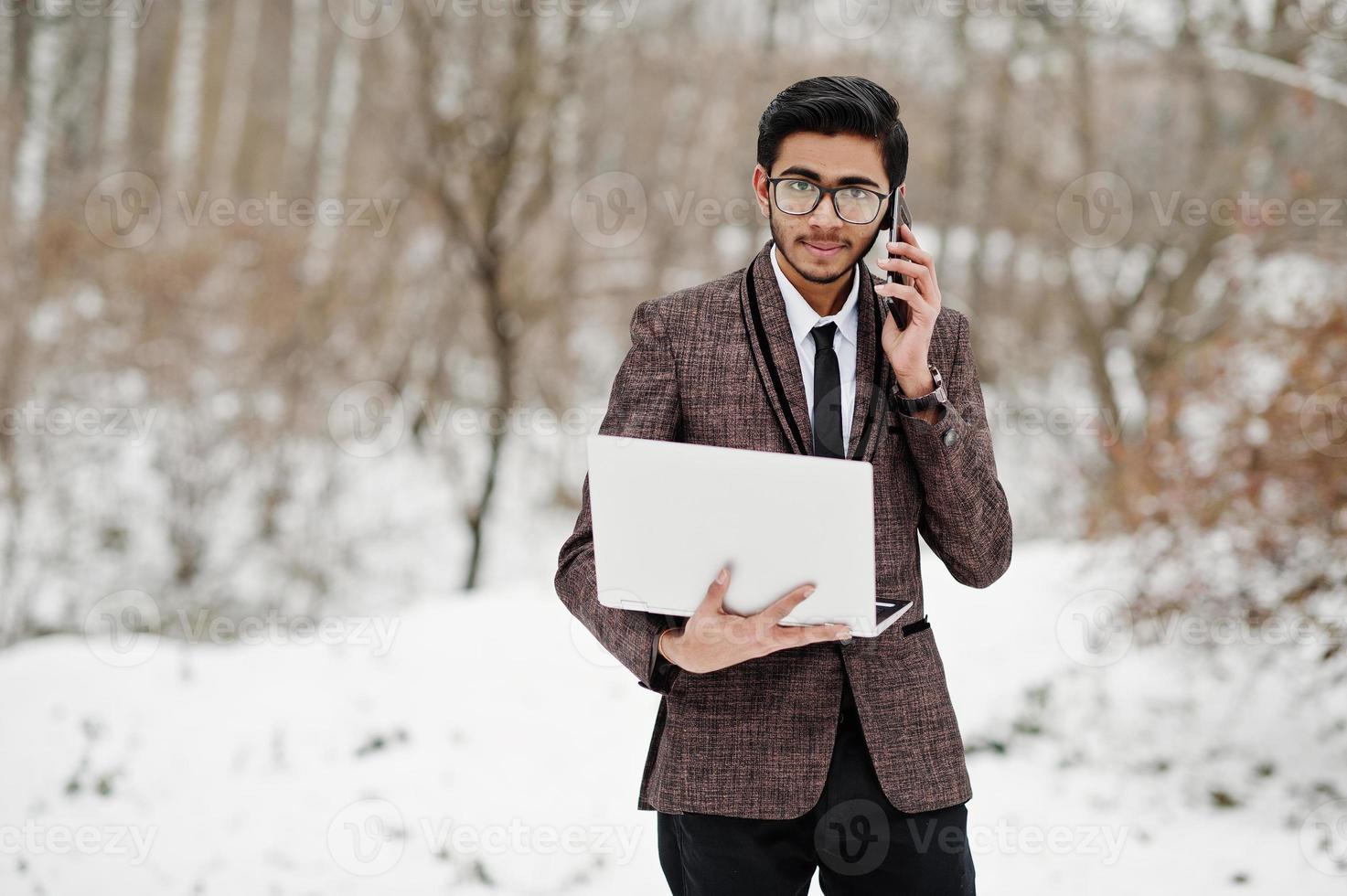 homme étudiant indien élégant en costume et lunettes posé à la journée d'hiver en plein air avec un ordinateur portable à la main, parlant au téléphone. photo