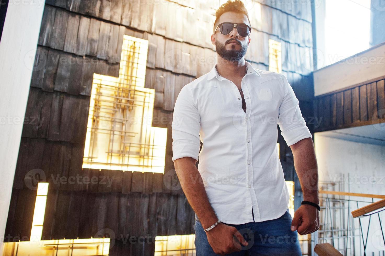 élégant grand modèle d'homme arabe en chemise blanche, jeans et lunettes de soleil posés contre un mur lumineux à l'intérieur. Barbe mec arabe attrayant. photo