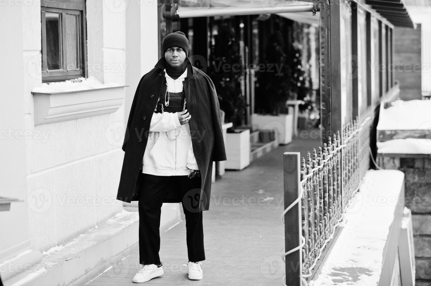 homme afro-américain élégant de style urbain en manteau noir et chapeau, sweat à capuche rose posé le jour de l'hiver. photo