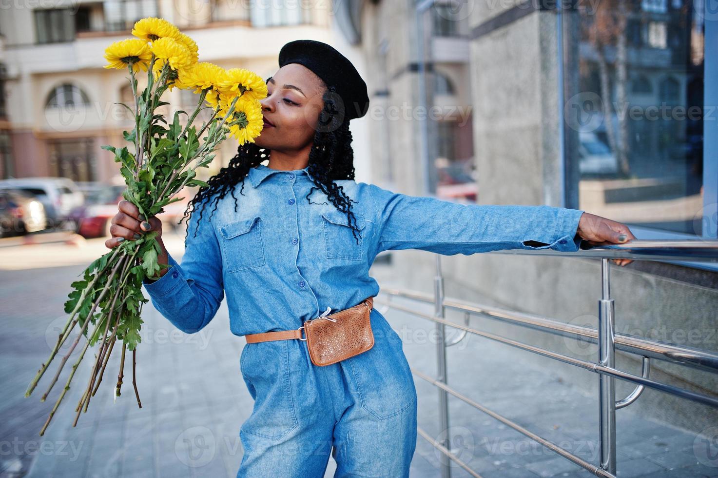 élégantes femmes afro-américaines à la mode en jeans et béret noir avec bouquet de fleurs jaunes posé en plein air par temps ensoleillé contre un bâtiment moderne bleu. photo