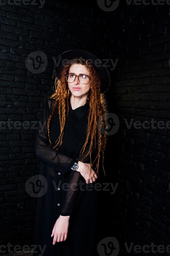 tournage en studio d'une fille en noir avec des dreads, des lunettes et un chapeau sur fond de briques. photo