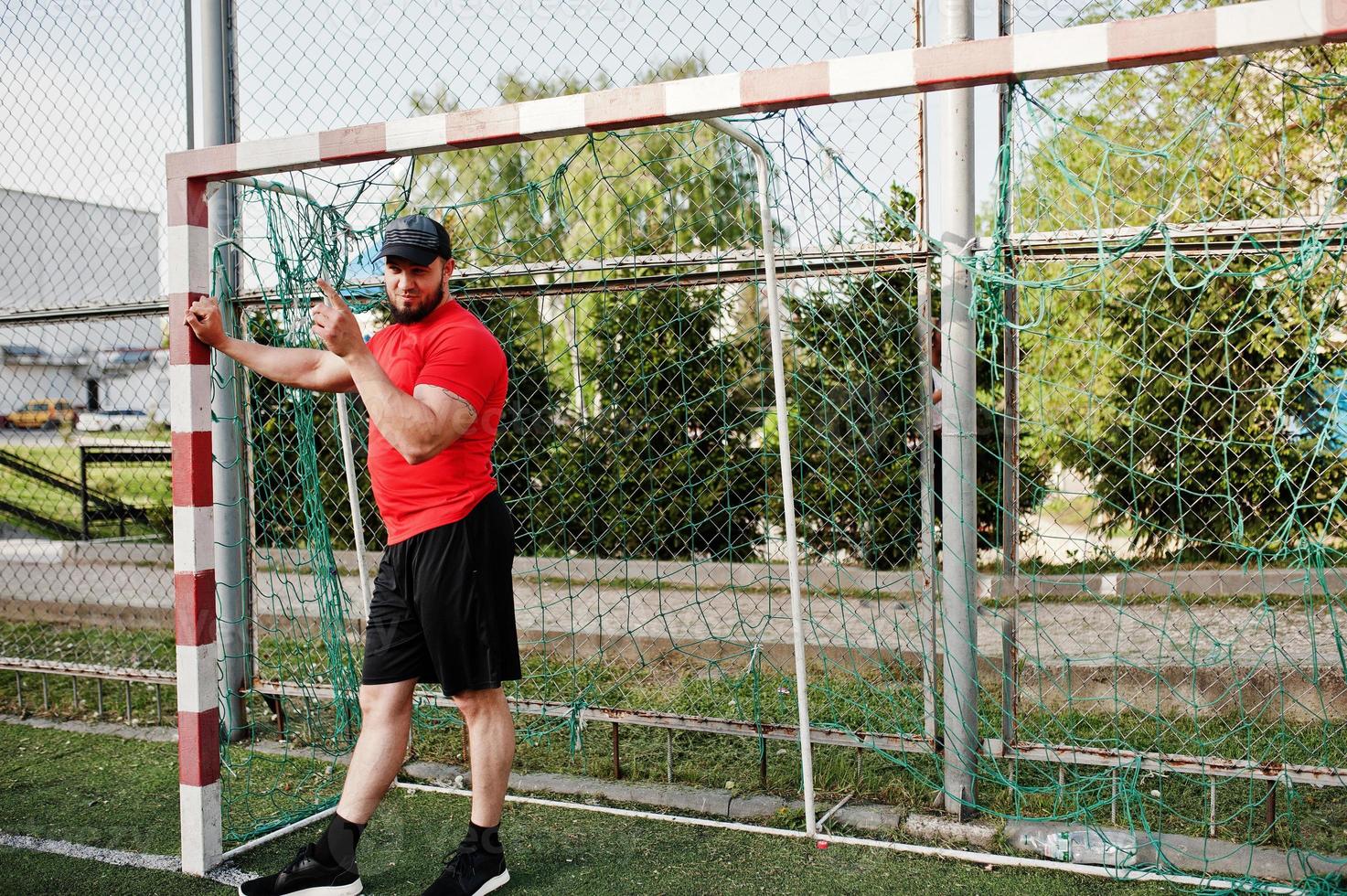 jeune homme musclé barbu brutal portant une chemise rouge, un short et une casquette au stade. photo