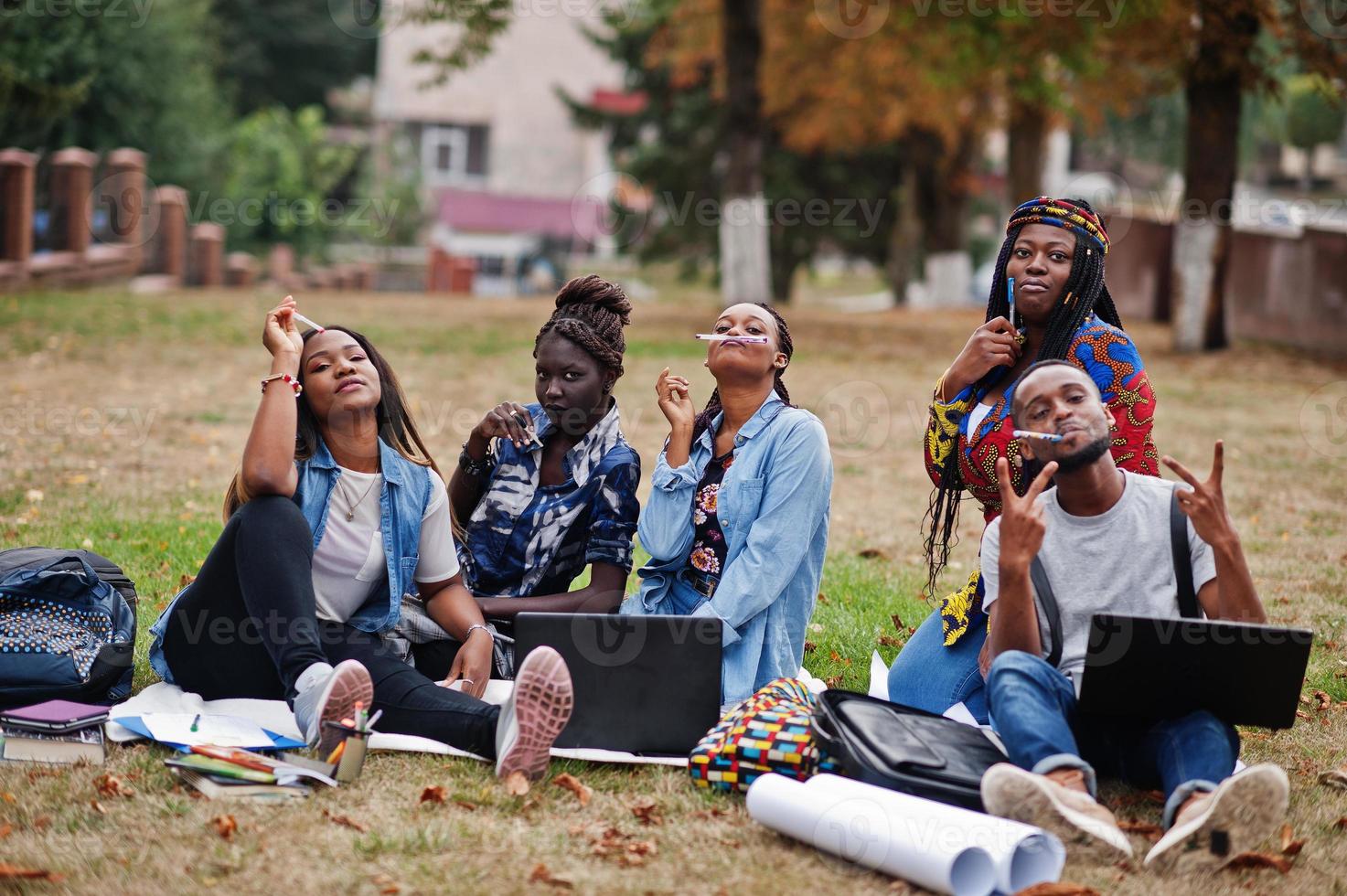 groupe de cinq étudiants africains qui passent du temps ensemble sur le campus de la cour universitaire. amis afro noirs assis sur l'herbe et étudiant avec des ordinateurs portables. photo