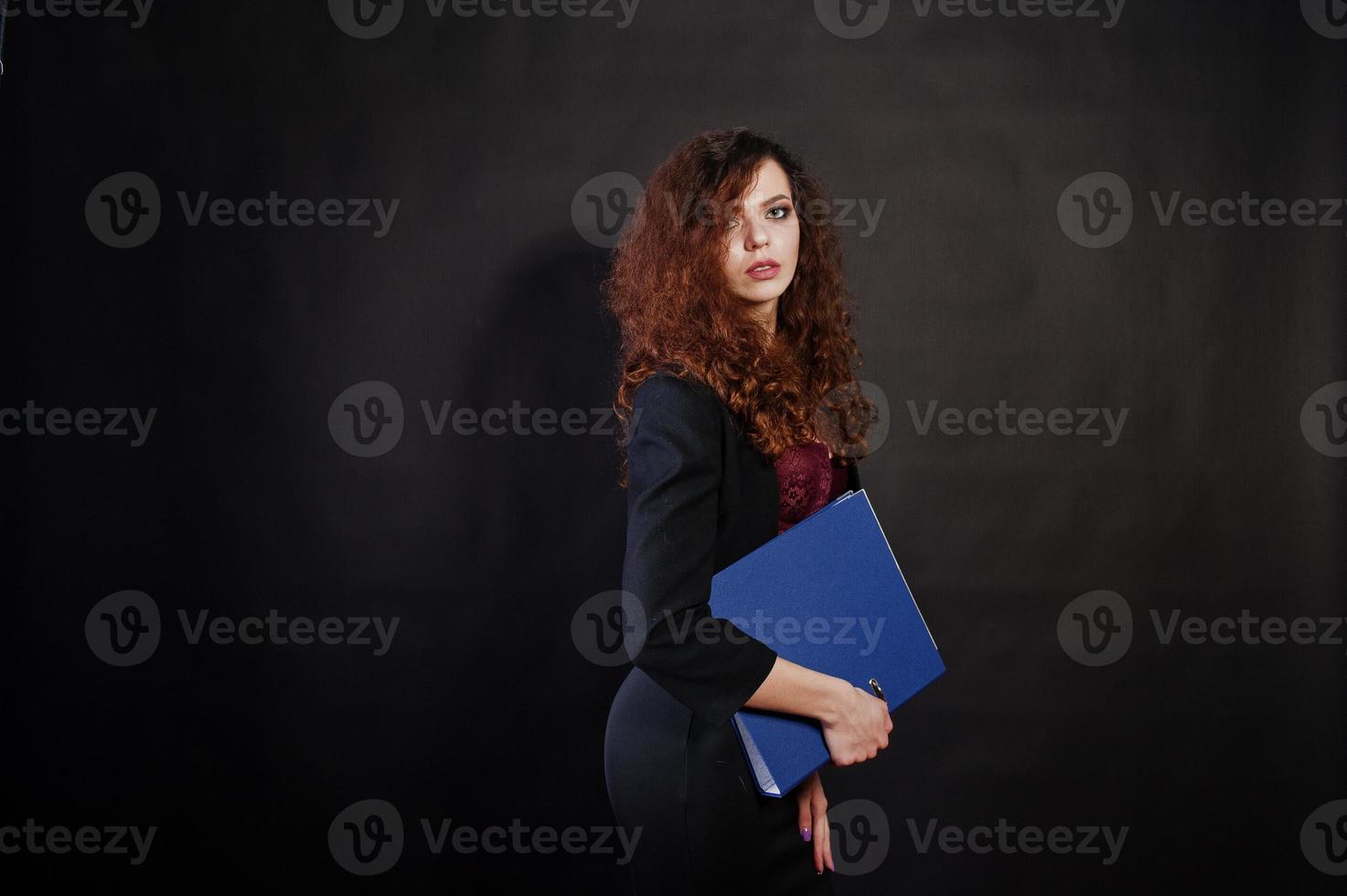 fille brune aux cheveux bouclés en veste de bureau noire avec jupe, sur soutien-gorge avec dossier de documents à mains sur studio sur fond noir. femme d'affaires sexy. photo