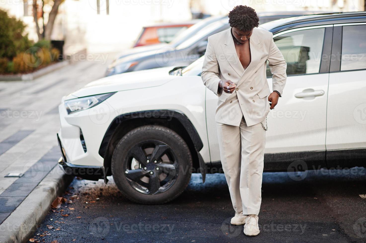 homme afro élégant en costume beige old school contre une voiture suv électro hybride moderne. jeune homme africain à la mode en veste décontractée sur torse nu. photo