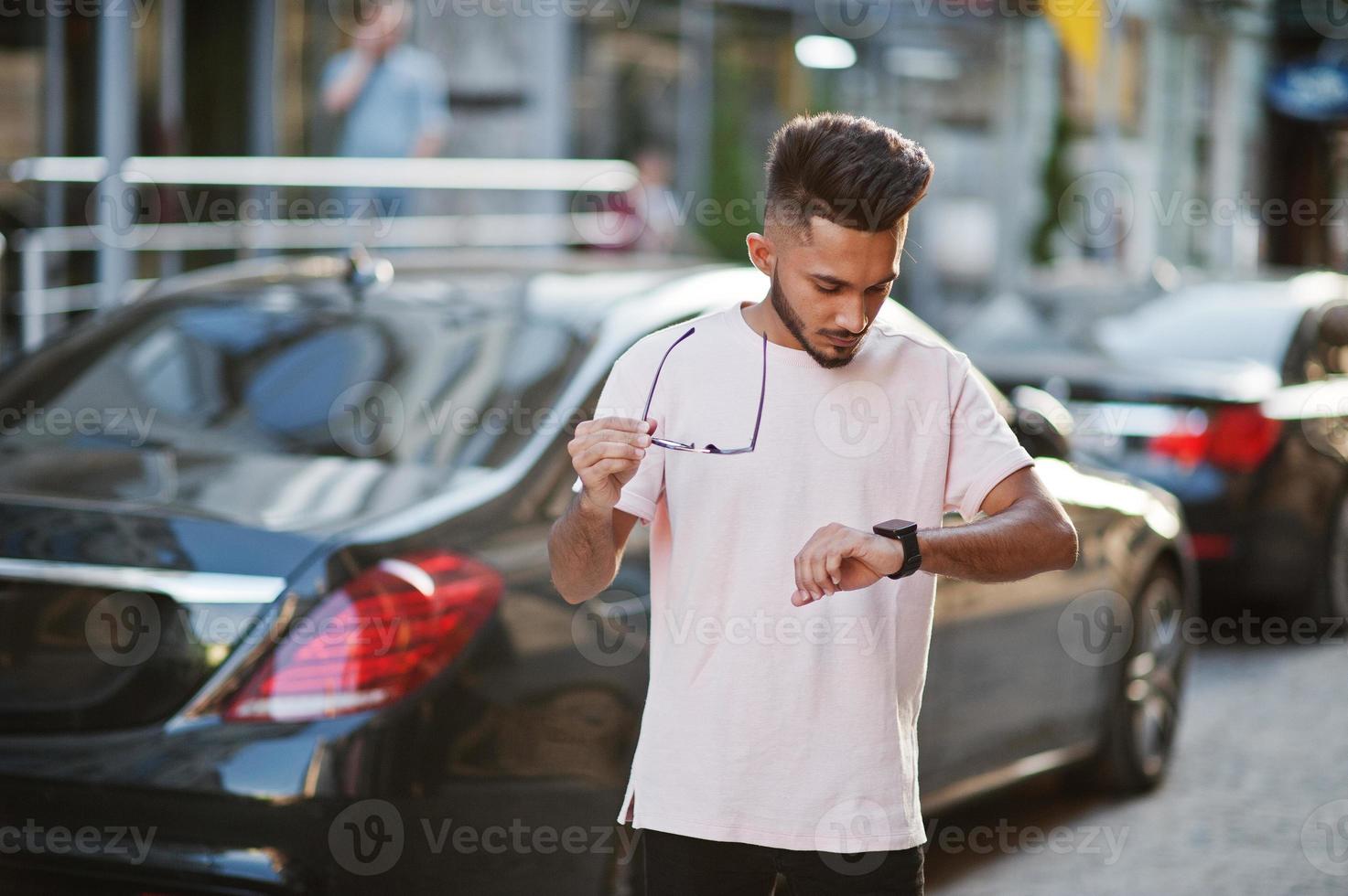 homme de barbe indien élégant à lunettes de soleil et t-shirt rose contre voiture de luxe. modèle riche indien posé en plein air dans les rues de la ville et regardant ses montres. photo