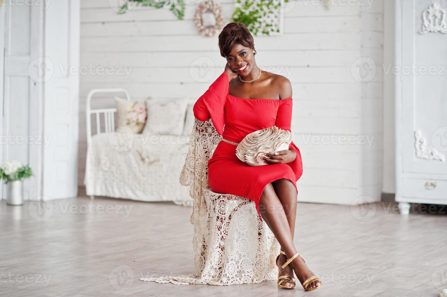 modèle afro-américain de mode en robe de beauté rouge, femme sexy posant une robe de soirée assise sur une chaise dans une salle vintage blanche. photo