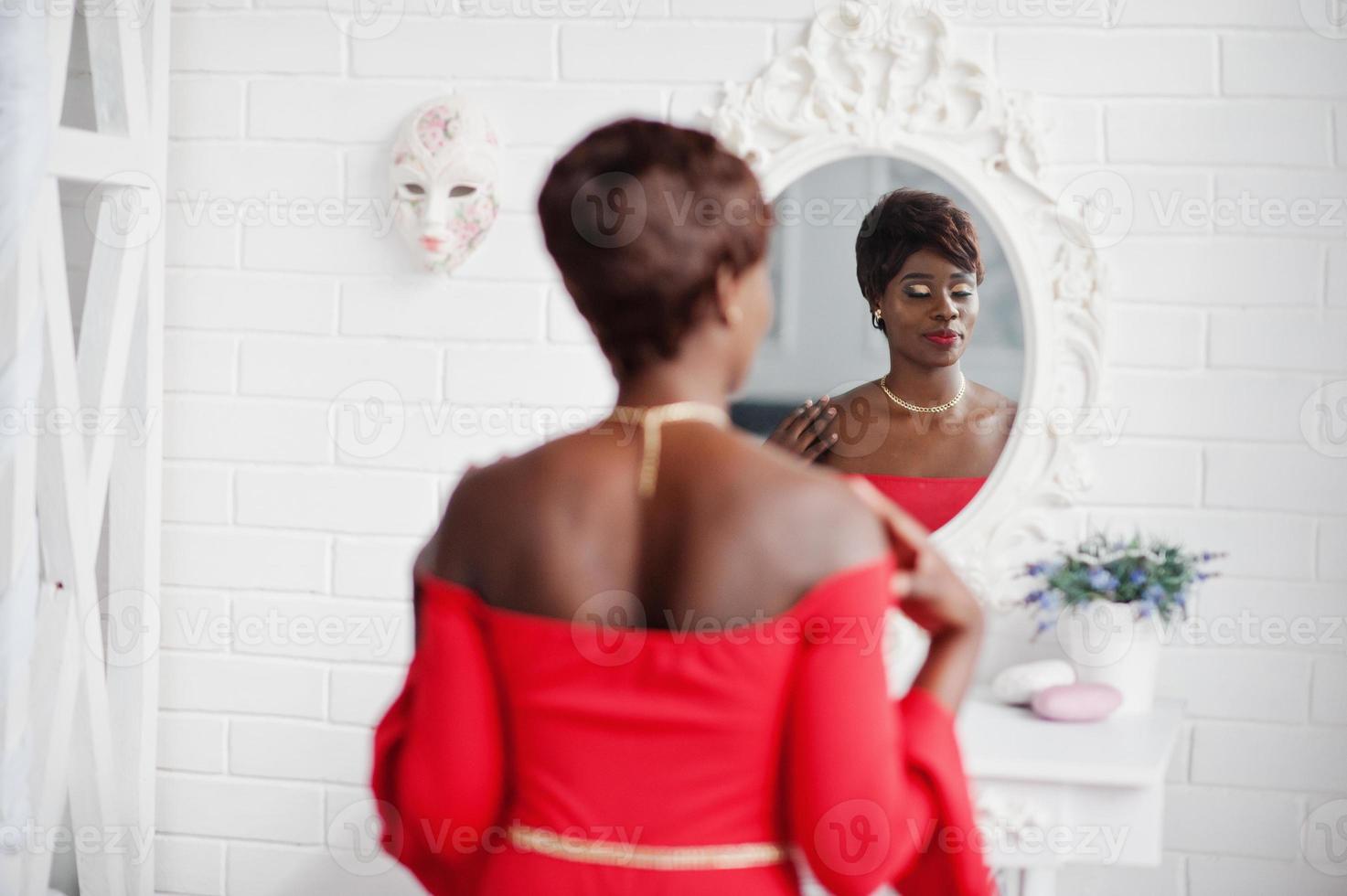 modèle afro-américain de mode en robe de beauté rouge, femme sexy posant une robe de soirée et regardant le miroir. photo