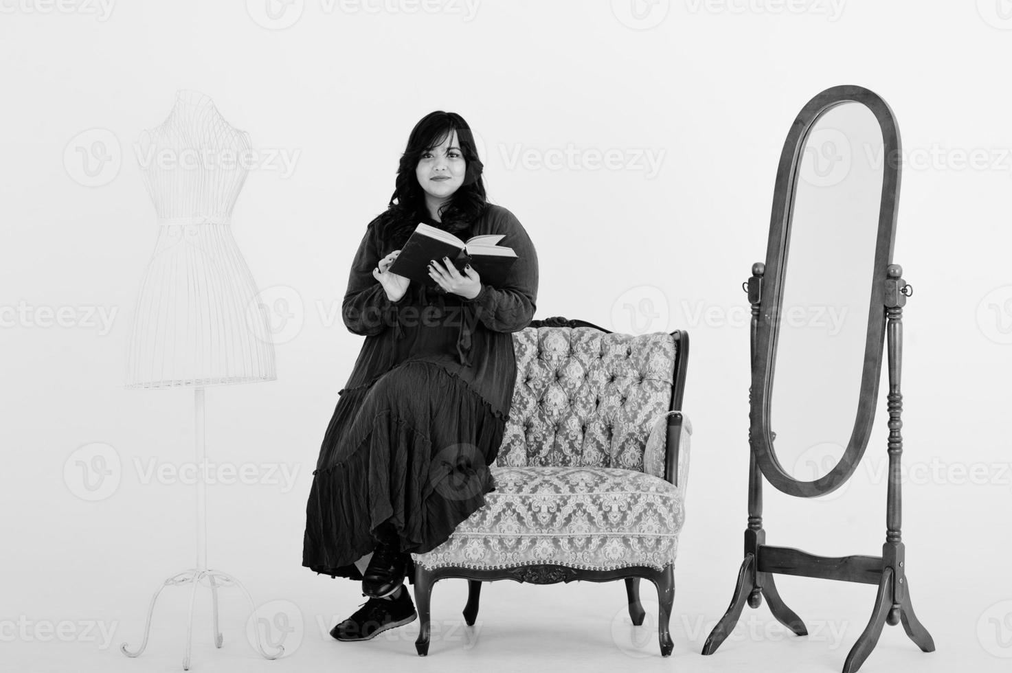 jolie femme sud-asiatique en robe rouge foncé posée au studio sur fond blanc contre le miroir et la chaise, lire un livre. photo