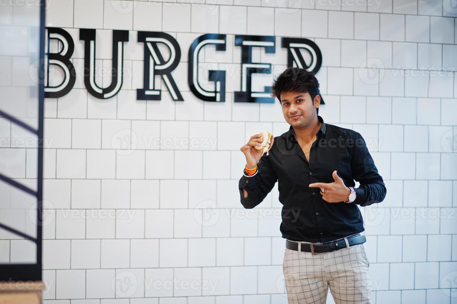 homme indien élégant au café de restauration rapide avec hamburger à portée de main contre le signe de burger sur le mur. photo