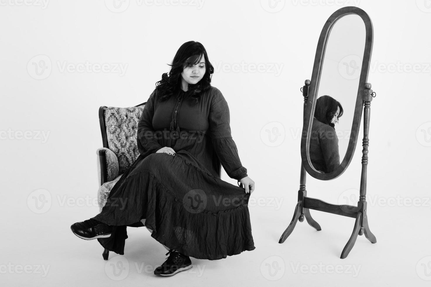 jolie femme sud-asiatique en robe rouge foncé posée au studio sur fond blanc contre le miroir et assis à la chaise. photo