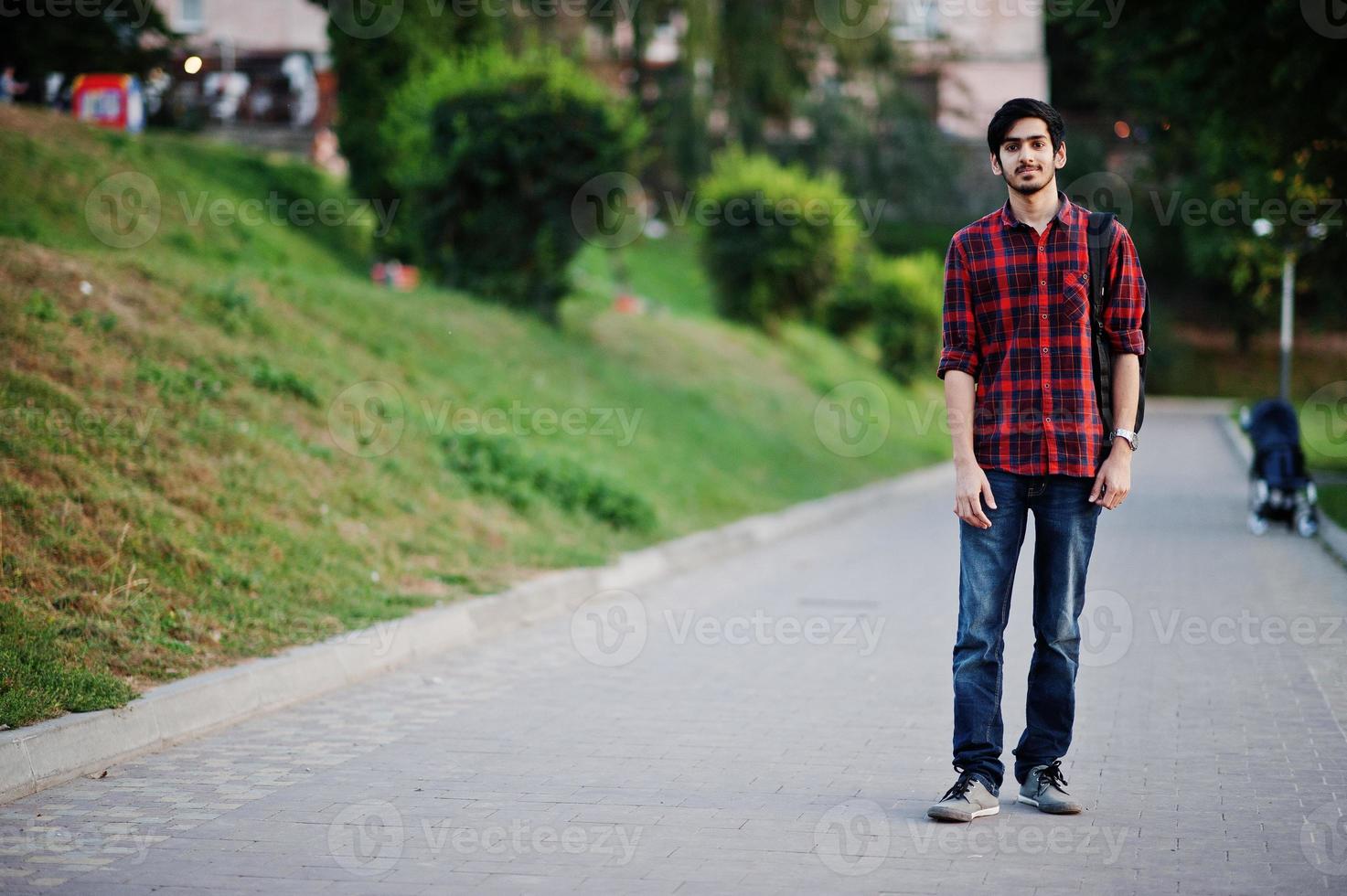 jeune étudiant indien en chemise à carreaux rouge et jeans avec sac à dos posé dans la rue. photo