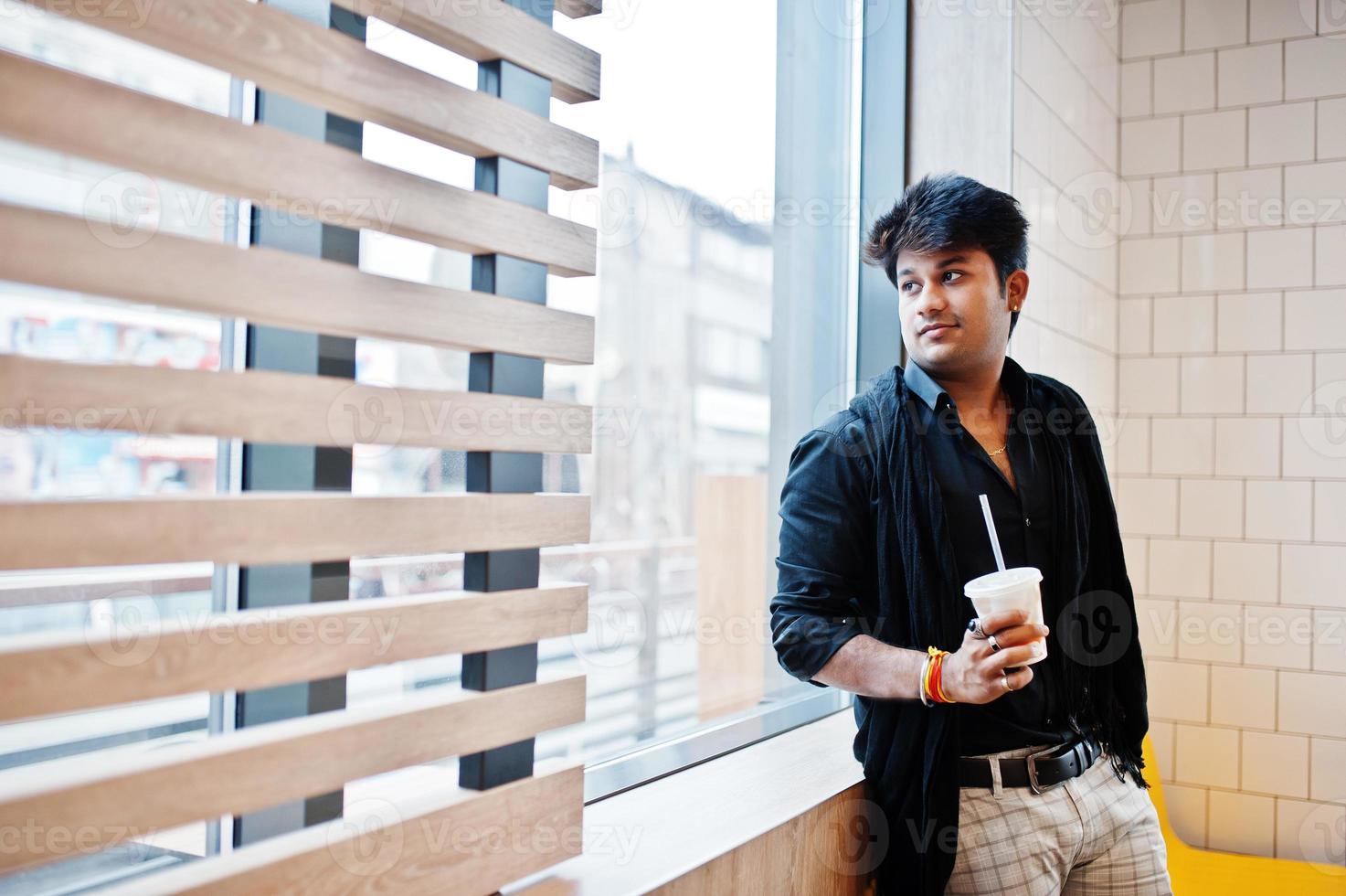 un homme indien élégant reste près de la fenêtre avec une tasse de café dans un café de restauration rapide. photo